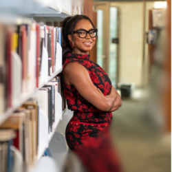 Abbey Fajimolu, owner of FFG Consulting, leaning against a bookshelf in a library in Mansfield, TX.